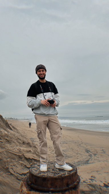 A young man wearing a beanie, a black and white jacket, and tan cargo pants stands on a wooden stump at the beach, holding a camera, with the ocean and cloudy sky in the background.