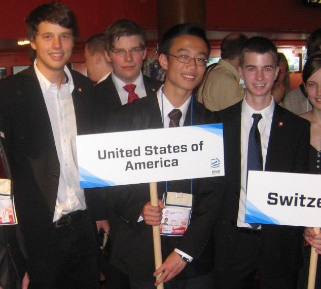Four young men in dark suits and ties stand together holding signs reading "United States of America" and "Switze[rland]," apparently representing their countries at what looks like an international conference or competition. One participant wears a lanyard with an "IPHO" (International Physics Olympiad) badge visible. The image illustrates the global, collaborative nature of academic competitions—a parallel to how international cooperation and shared intellectual rigor contribute to advancing technical fields, much like the investment in HPC referenced in the post about scaling Hyperliquid's development.