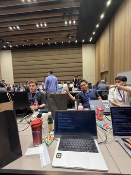 A conference or hackathon setting with multiple people working on laptops at long tables in a large hall.