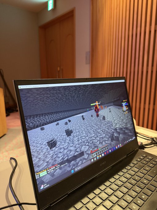 The image shows an open laptop computer displaying a Minecraft game. The screen of the laptop displays a map of a virtual world filled with various objects such as blocks and other Minecraft elements. The laptop is placed on top of a table, which also has some cables connected to it. In the background, there are two doors visible behind the laptop.