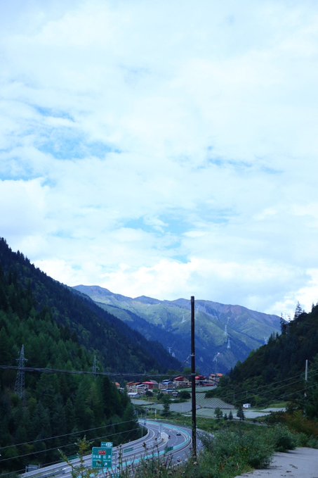A scenic mountain landscape featuring a winding road through a valley with small houses, forested hills, and power lines under a cloudy blue sky.