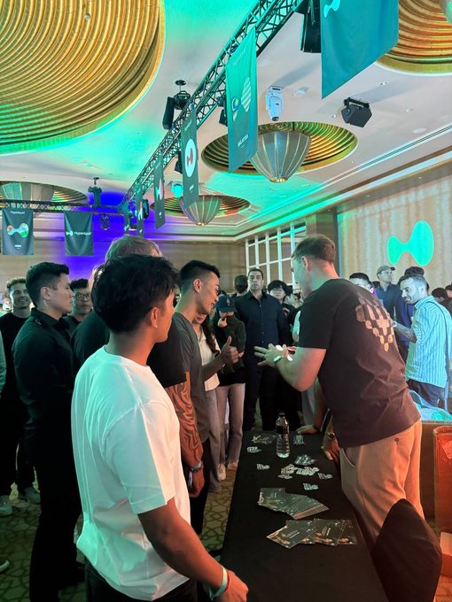 A crowd of people gathers around a table at what appears to be a product demonstration or promotional event, with a woman on a phone and a presenter addressing attendees under green lighting and decorative ceiling fixtures.