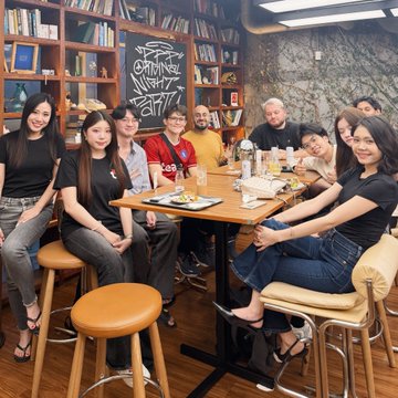 A group of nine people pose together around a wooden table in a cafe with bookshelves and graffiti-decorated walls in the background.