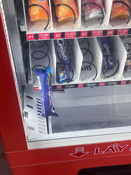 A vending machine with prices in VND showing snacks and drinks on numbered shelves (41-55). An Oreo ice cream bar is stuck/half-dispensed from slot 51, and the machine displays a 'LẤY' (Take) label with a Vietnamese language indicator.