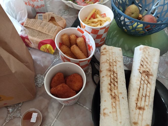 A spread of fast food on a table including two grilled burritos/wraps, McDonald's nuggets and fries in red checkered cups, a hash brown, a fruit basket, and a brown paper bag.