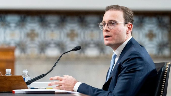 A man in a dark suit and glasses speaking at a formal hearing or testimony with a microphone, water bottles, and documents on the table before him.