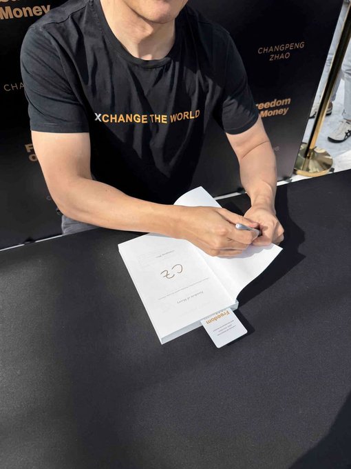 A man wearing a black "XCHANGE THE WORLD" t-shirt sits at a book signing table, signing a copy of his book as a backdrop displays "CHANGPENG ZHAO" and "Freedom Money." The image reflects the post's theme of building toward a vision, capturing CZ—founder of the Binance "Xchange"—sharing his book about financial freedom with supporters.