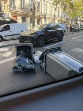 A black SUV is stopped on a Parisian street next to two overturned metallic trash bins with scattered black garbage bags spilling onto the road, with a white van and a motorcyclist visible in the background.