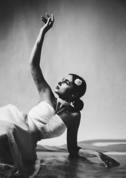 A black-and-white photograph captures a dancer in a white gown kneeling with one arm extended gracefully upward, a white flower adorning her dark hair.