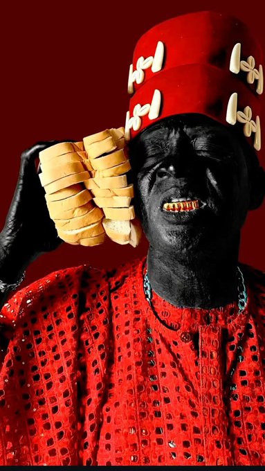 A person with dark skin wearing an ornate red hat and matching red embroidered robe, smiling to reveal gold teeth while holding a large yellow braided object.