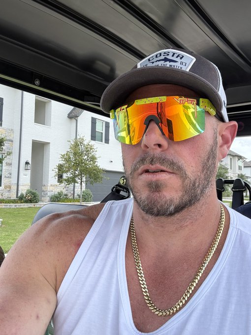 "Mog coin" is a term used for a gold chain that has been designed by the artist, Olafur Eliasson. It features a unique design of a tree with branches made from metal wires. The image shows a man wearing a white tank top and sunglasses, possibly in a car or on a golf cart, which could be an advertisement for the "Mog coin" chain.