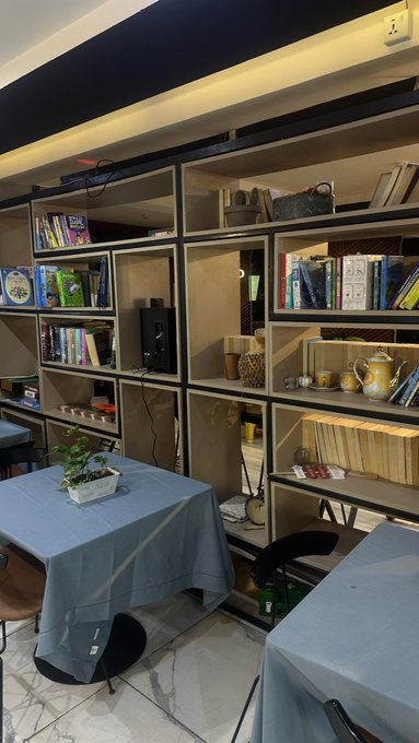 Interior view of a room featuring wooden bookshelves filled with books and decorative items, with a table covered in a light blue tablecloth and chairs around it.