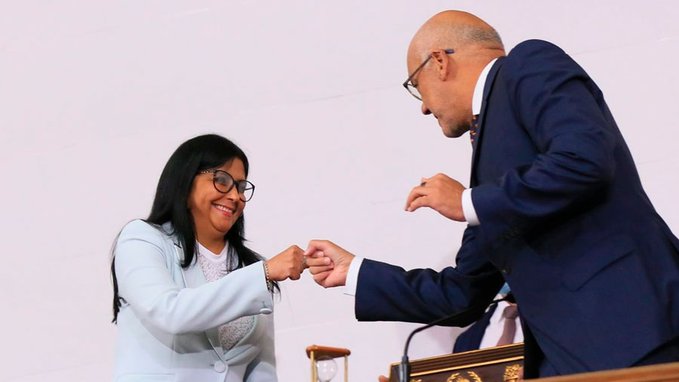 A woman in a light blue blazer smiles warmly as she fist-bumps a bald man in a dark suit wearing glasses.