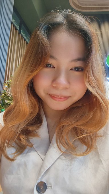 A young woman with long, wavy caramel-highlighted hair smiles softly in a cozy selfie, wearing a cream-colored coat against a warm, decorated indoor backdrop. Her fresh-faced glow suggests she's embracing the weekend's relaxation, perfectly matching the "Happy Sunday" vibes of enjoying some downtime.