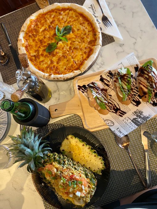 A table is set with a pizza, a wooden board of bruschetta drizzled with balsamic, and a hollowed-out pineapple dish filled with a colorful salad.