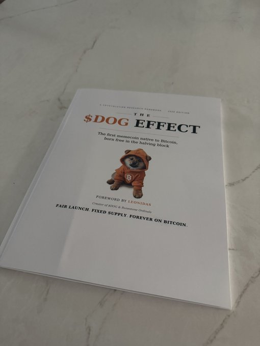 A copy of "The $DOG Effect" book rests on a marble surface, featuring a Shiba Inu in an orange hoodie on the cover. The book is described as "The first memecoin native to Bitcoin, born live in the halving block" with a foreword by Leonidas, and its tagline reads "FAIR LAUNCH. FIXED SUPPLY. FOREVER ON BITCOIN."