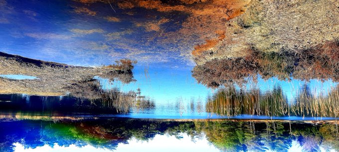 An artistically mirrored landscape photo showing a rocky hillside, trees with orange autumn foliage, blue sky, and water, with the reflection creating a symmetrical abstract effect.