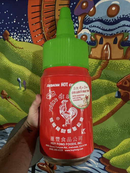 A hand holds a red Sriracha Hot Chili sauce bottle with a green cap, featuring a 'Stray Kids Collectible Tin with Chopsticks and Salted Popcorn' promotional sticker, set against a colorful illustrated background.