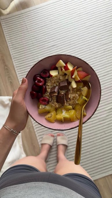 The image features a person standing next to a pink bowl filled with fruit, including cherries and apples. The person appears to be holding the bowl up for the camera, showcasing the colorful assortment of fruits inside. The background includes a rug on which the person is standing, as well as some text that reads "GM CT starting sunday a little slower today hope you all have a peaceful day ahead".