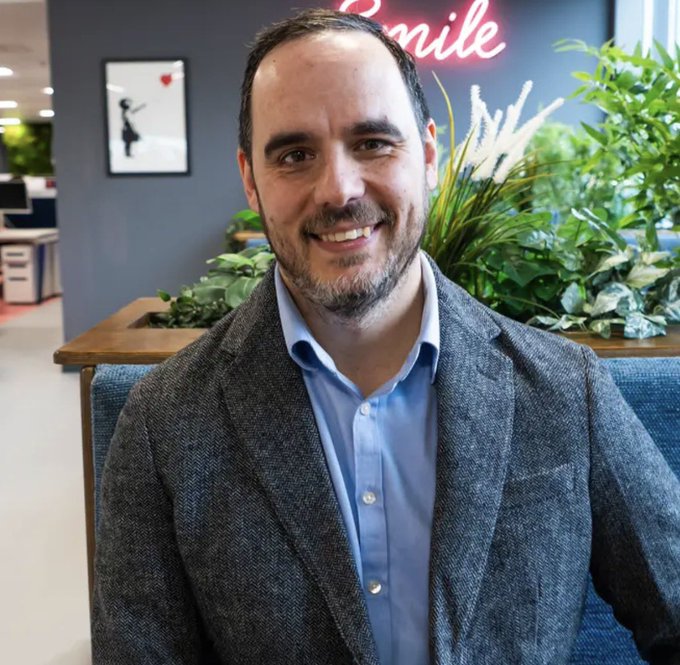A smiling man with a beard wearing a grey blazer over a light blue shirt, seated in a modern office with greenery and a neon 'Smile' sign in the background.