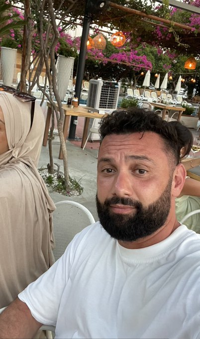 A bearded man in a white t-shirt takes a selfie at an outdoor restaurant patio decorated with vibrant pink bougainvillea, while a woman wearing a beige hijab sits beside him under the pergola. The image captures his casual dining outing as part of his "Day 4" social media campaign advocating for the $HYPE cryptocurrency token to reach a 3-digit price.
