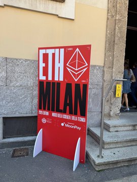 irt's ETH Milan sign, featuring a red and white logo with a black triangle, stands on a sidewalk next to a building entrance.