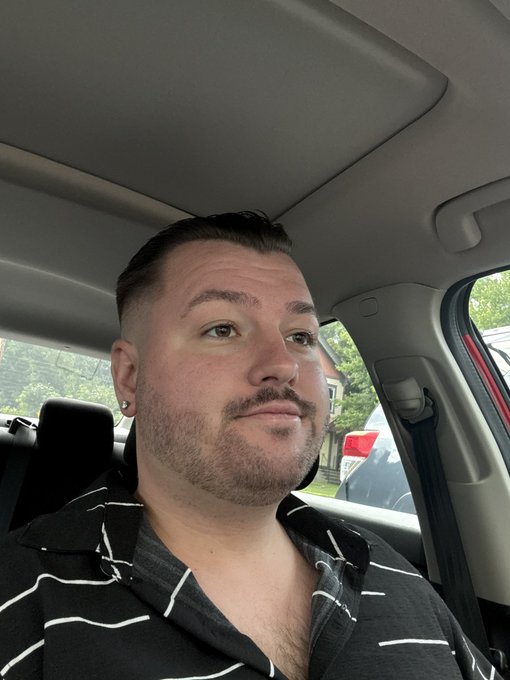 A selfie of a man with a short beard and dark hair sitting in a car, wearing a black and white striped shirt.