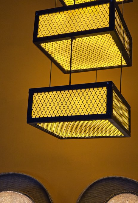 Two geometric, box-shaped light fixtures with bright yellow mesh-paneled panels hang suspended from a warm ochre ceiling, their vibrant glow complementing the post's "Yellow" theme.