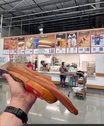 A first-person view of a hand holding an exceptionally long hot dog in a bun inside a warehouse store, with food court menu boards visible in the background.