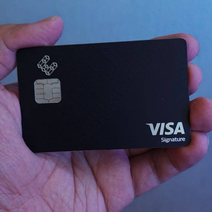 A hand holds a black Visa Signature credit card featuring a metallic chip and a geometric logo at the top.
