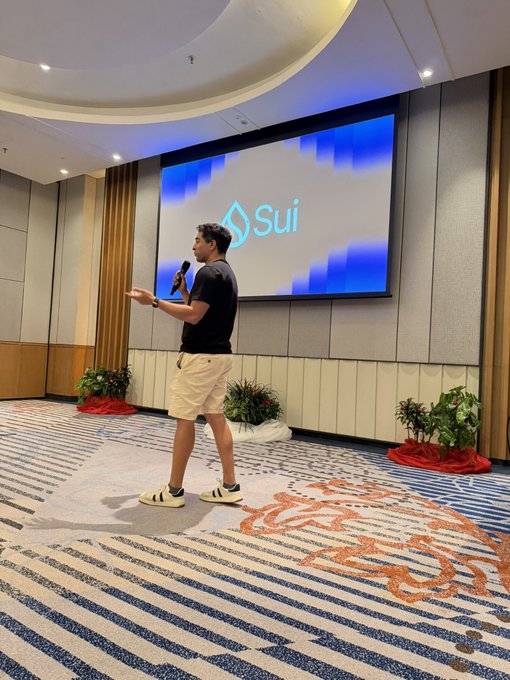A man stands on a patterned carpet speaking into a microphone, presenting in front of a large screen displaying the 'Sui' logo.