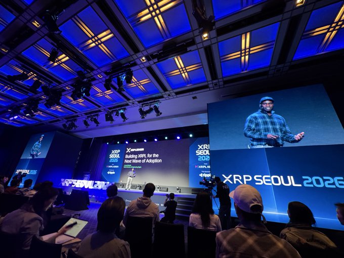 A speaker presents on stage at the XRP Seoul 2026 conference, with his image displayed on a large screen to the audience seated in a dimly lit auditorium.
