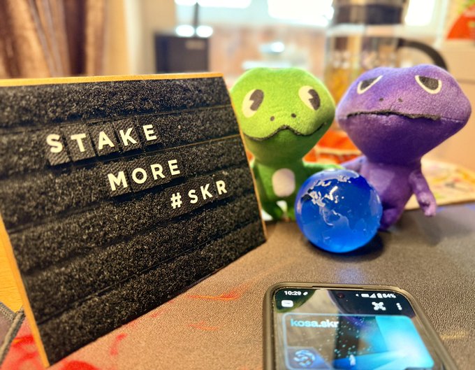 A photo showing a letterboard reading 'STAKE MORE #SKR' next to two small plush dinosaur toys (green and purple), a blue glass globe, and a smartphone on a table.