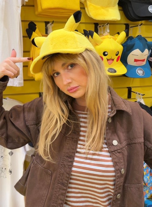 A blonde woman wearing a yellow Pikachu hat and brown jacket points at herself in a store, with Pikachu and Snorlax plush hats displayed on a yellow wall behind her.