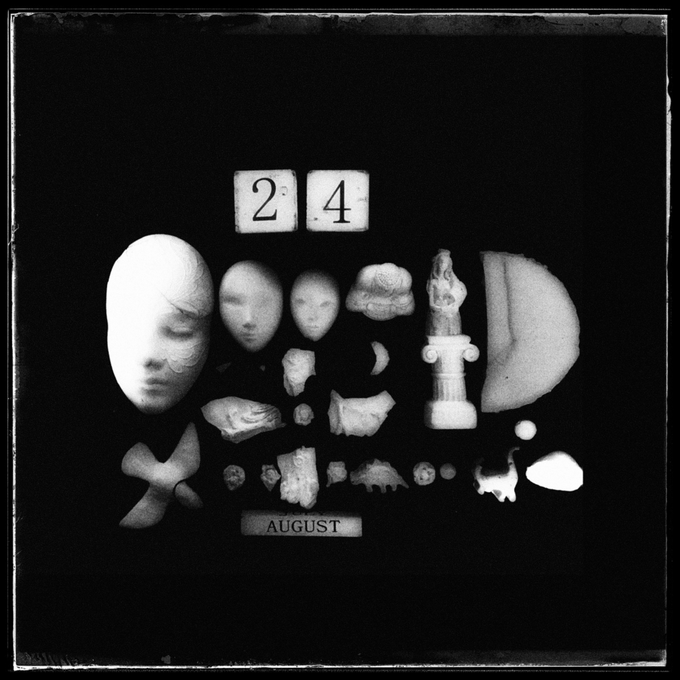 Dark, artistic black and white photograph from 1917 showing various sculptural fragments including face masks and pieces arranged with calendar blocks displaying '24' and a label reading 'AUGUST'.