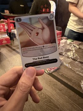 A hand holds up a custom card titled "The Red Bean" featuring artwork of a red bean in a hand, appearing to be from a TCG-style game.