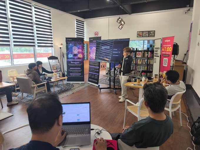 The image shows a group of people gathered around a table with laptops open, possibly working together or attending an event. There are at least five individuals visible in the scene, including one person standing near a large screen displaying information about Bitcoin. The room has white walls and black blinds that allow natural light to enter. A few chairs can be seen scattered throughout the space, along with a dining table located towards the back of the room.