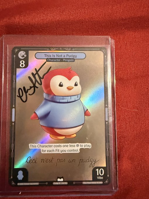 A holographic 'This Is Not a Pudgy' trading card featuring a penguin character, signed in black marker, encased in a protective sleeve on a red surface.