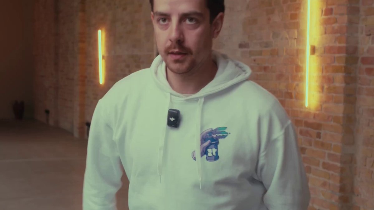 The image features a man wearing a white hoodie with an emblem of a dragon design. He has a black cell phone attached to his jacket and appears to be looking off into the distance, possibly focusing on something or someone outside the frame of the picture. The surrounding environment includes yellow lights that illuminate the scene, adding a sense of atmosphere and intrigue to the photo.