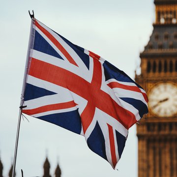A Union Jack flag waves prominently in the foreground with the iconic Big Ben clock tower visible in the blurred background, symbolizing the United Kingdom as Robinhood expands its crypto trading services to the British market.