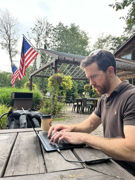 A man in a brown shirt works on a laptop at an outdoor wooden table with an American flag visible in the background.