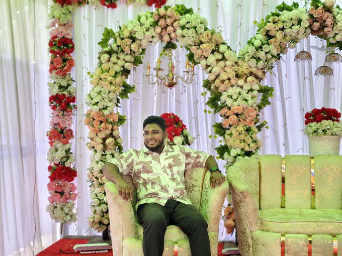 The image features a man sitting on a chair with his legs crossed, wearing a white shirt. The background of the photo shows an elaborate floral arrangement behind him, which includes red, pink, green, and white flowers. There are also some books visible in the scene. The caption to the image reads "Good Morning X fam! No noise, just pure building.Bullish on what @XOOBNetwork and @useTria are bringing to the table." This suggests that the man is sitting in a room with a beautiful floral arrangement as decoration while reading some books.
