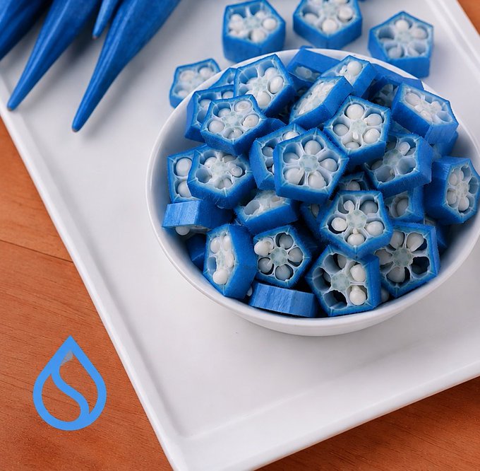 The image features a white plate with blue food items arranged on top of it. The food consists of small blue pieces that appear to be cut in half or quarters. There are also some blue fruits, possibly okra, placed around the plate. The background is blurred and does not provide any additional information about the context of the scene.