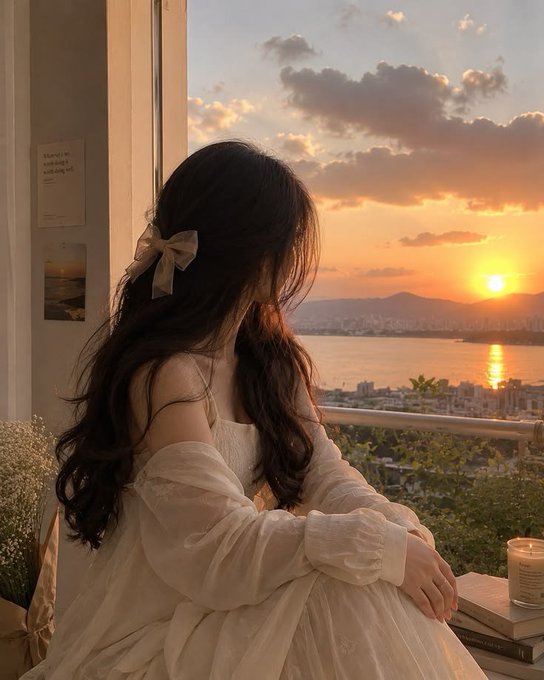 A woman with long dark hair wearing a white off-the-shoulder dress looks out from a balcony at a sunset over a coastal city.