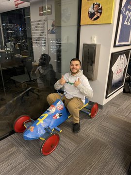 A photograph of a smiling man in a white hoodie and khaki pants sitting on a blue toy airplane chair on wheels inside an office with framed sports jerseys on the walls.