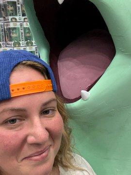 A selfie of a woman with reddish hair wearing a backwards blue and orange baseball cap, posing next to a large green cartoon creature with an open mouth and visible tooth.