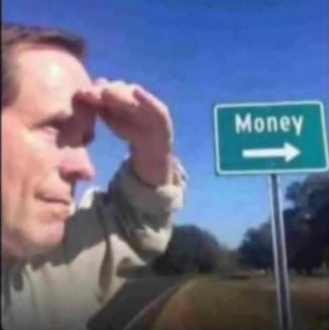 A man shielding his eyes as he looks into the distance, with a green road sign beside him pointing right with the word 'Money' written on it.