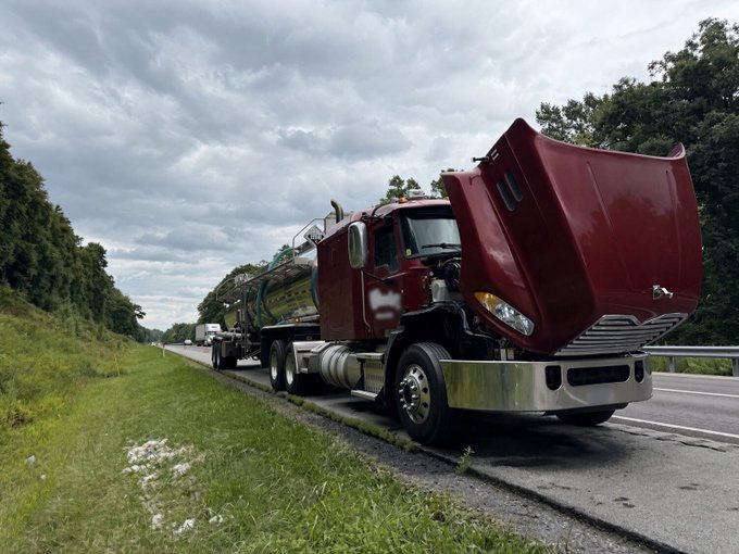 A photograph of a damaged maroon semi-truck on the shoulder of a highway, with its hood ajar and debris visible on the road beneath it, under a cloudy sky.