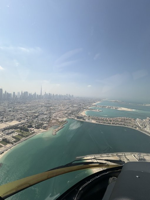 An aerial view from a cockpit window shows Dubai's coastline with the Burj Khalifa skyline in the distance and the iconic Palm Jumeirah visible on the right, captured mid-flight as a sunlit "GM" greeting for the day ahead.