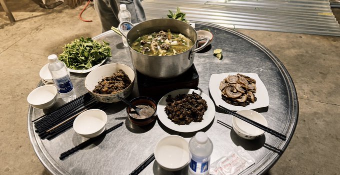 A round metal table set up for a communal meal outdoors, featuring a large pot of chicken soup, plates of greens, mushrooms, and other dishes surrounded by chopsticks and water bottles.